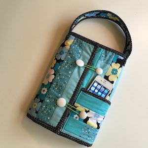 May include: A quilted fabric bag with a black and white polka dot trim. The bag has a blue, yellow, and white floral pattern and features a pocket for a phone. The bag has a handle and two snap closures.