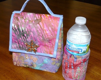 May include: A quilted lunch bag with a blue handle and a pink and purple floral pattern. The bag has a small jeweled flower brooch on the front. A clear plastic water bottle with a blue and white label that says "Ice Mountain" is in a matching quilted bottle holder.