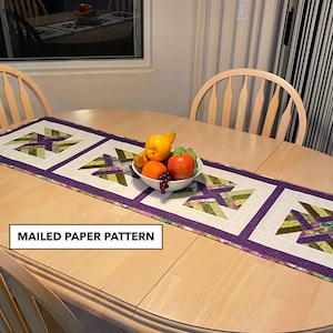 May include: A table runner with a purple border and a white background featuring a green and brown geometric pattern. The runner is on a wooden table with a bowl of fruit in the center. The text "MAILED PAPER PATTERN" is visible in the lower left corner of the image.