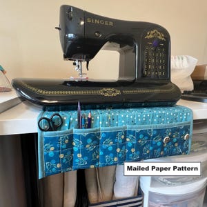May include: A blue and white quilted sewing machine organizer with pockets for storing sewing supplies. The organizer is hanging on a white table under a black Singer sewing machine. Mailed Paper Pattern.