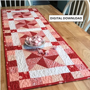 May include: A red and pink patchwork table runner with a white background. The runner is quilted with a swirl pattern and has a red border. The runner is on a wooden table with a bowl of sweets on top.