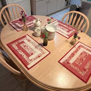 Op de afbeelding: Een lichthouten eettafel met vier rechthoekige stoffen placemats met een rood en crème bloemenpatroon. De tafel bevat een stapel witte keramische kommen, verschillende witte mokken en drie kleine glazen vazen met roze bloemen.