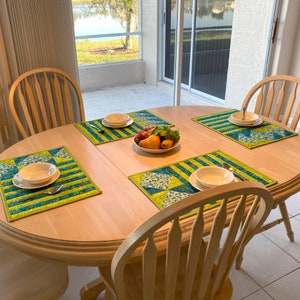 May include: Four placemats with a blue and green geometric pattern are set on a light wood table. A bowl of fruit sits in the centre of the table.
