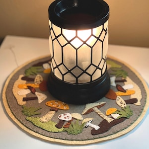 May include: A black and white wax warmer with a hexagonal design sits on a grey felt mushroom-themed coaster. The warmer is lit and glowing.