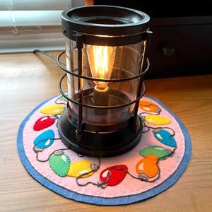 May include: A black metal lantern-style lamp with a clear glass enclosure and a lit Edison bulb. The lamp sits on a round pink felt coaster decorated with colorful felt Christmas lights. The coaster has a blue border.