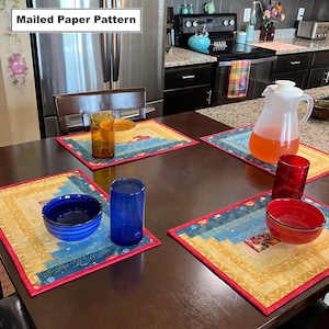 May include: Four placemats with a patchwork design in shades of blue, yellow, and red, with a red border. The placemats are on a dark brown wooden table with two blue glasses, two blue bowls, a clear glass with a brown liquid, and a red glass with a clear liquid. The text "Mailed Paper Pattern" is visible in the top left corner of the image.