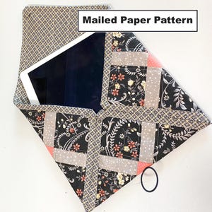 May include: A black and white fabric envelope with a floral pattern and a pink accent. The envelope is folded to create a pocket for a tablet or other device. The text "Mailed Paper Pattern" is printed on the envelope.