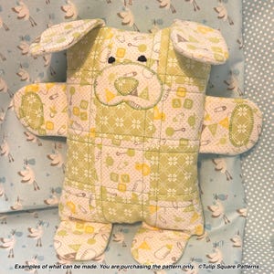 May include: A stuffed animal toy shaped like a dog, constructed from quilted fabric featuring a light green and white pattern of safety pins, baby blocks, and rattles. The toy has two floppy ears, two arms, and two legs. Text at the bottom reads: Examples of what can be made. You are purchasing the pattern only. ©Tulip Square Patterns.