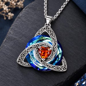 May include: A silver-toned pendant necklace featuring a triquetra knot frame surrounding a circular, multi-faceted blue and green crystal. At the centre of the crystal sits a small, vibrant orange heart-shaped gemstone, suspended from a silver chain.