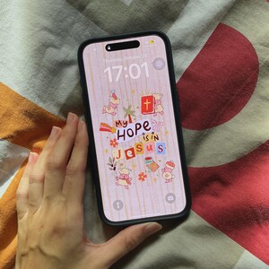 Op de afbeelding: Een zwarte smartphone met een scherm waarop de tekst "My Hope is in Jesus" in een kleurrijk, grillig ontwerp wordt weergegeven. Het scherm van de telefoon toont ook de datum en tijd. De telefoon wordt door een persoon vastgehouden.