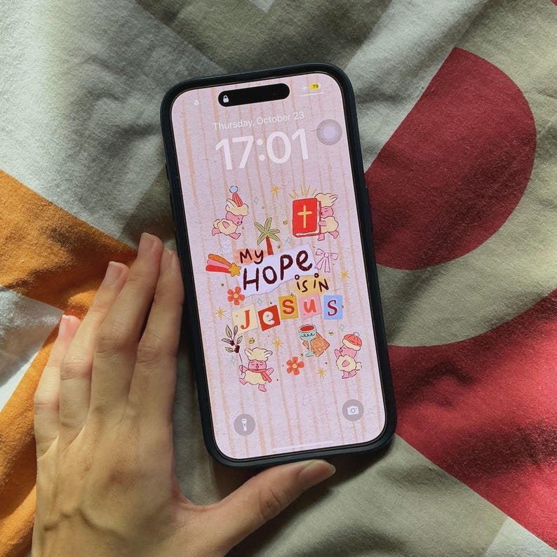 Op de afbeelding: Een zwarte smartphone met een scherm waarop de tekst "My Hope is in Jesus" in een kleurrijk, grillig ontwerp wordt weergegeven. Het scherm van de telefoon toont ook de datum en tijd. De telefoon wordt door een persoon vastgehouden.