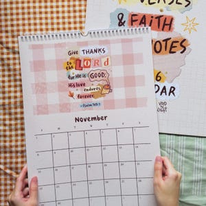 Può includere: Un calendario aperto a novembre, con un layout a griglia ed elementi decorativi. La parte superiore include il testo "Give Thanks to the Lord" con un'illustrazione di tazza di caffè. Una seconda pagina mostra le parole "Verses & Faith".