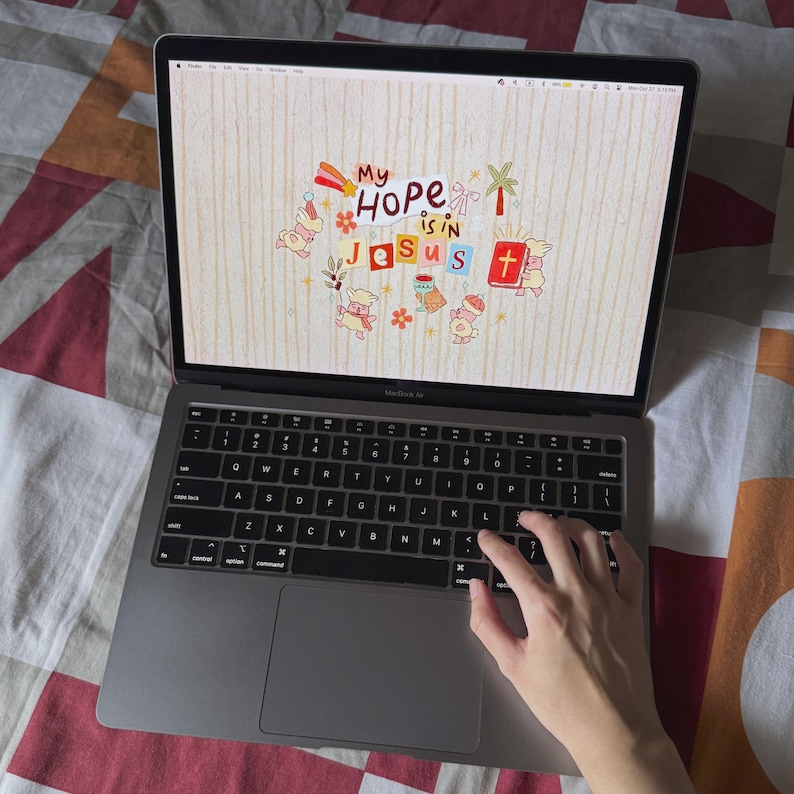 Op de afbeelding: Een zilveren MacBook Air laptop met een kleurrijke afbeelding met de tekst "My HOPE is in Jesus." Het zwarte toetsenbord is zichtbaar, met een hand boven de toetsen. De laptop staat open op een bed met een patroon.