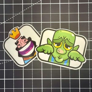 May include: Two stickers featuring characters from the game Clash Royale. One sticker shows a laughing king with a gold crown. The other sticker depicts a green goblin with a sad expression and blue overalls.
