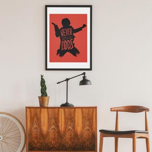 May include: Black framed art print featuring a silhouette of a figure holding a blaster, set against a red background. The text "NEVER tell me the ODDS" is displayed in black. The print is displayed above a wooden cabinet.