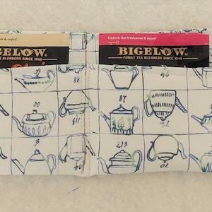 Puede incluir: Un soporte de tela para bolsitas de té con cuatro bolsillos, cada uno con una bolsita de té Bigelow. El soporte es blanco con un estampado de tetera azul. Una correa negra está unida al lateral. Las bolsitas de té están etiquetadas con la marca Bigelow.