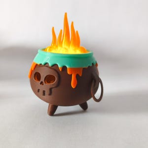 May include: A brown cauldron with a skull design, teal rim, and orange drips, topped with orange and yellow flames. The cauldron has three small legs and a metal ring handle. A whimsical, Halloween-themed decoration.