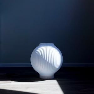 May include: A white, ribbed, spherical vase with a rounded base. The vase is illuminated by sunlight, casting a shadow on the wooden floor. The background is a solid, dark blue wall.