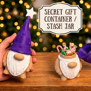 Op de afbeelding: Een kaboutervormige houder met een afneembare paarse muts met een witte ster. Het lichaam is beige met een witte baard en neus. Eén pot is geopend en bevat snoepjes. De tekst luidt: SECRET GIFT CONTAINER / STASH JAR.