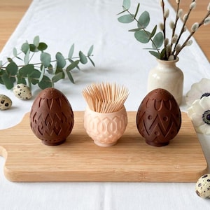 Op de afbeelding: Paasdecoratie met twee donkerbruine eivormige zout- en peperstrooiers met decoratieve patronen, een lichtroze tandenstokerhouder en kwarteleieren op een houten plank. Eucalyptus en bloemen maken het af.
