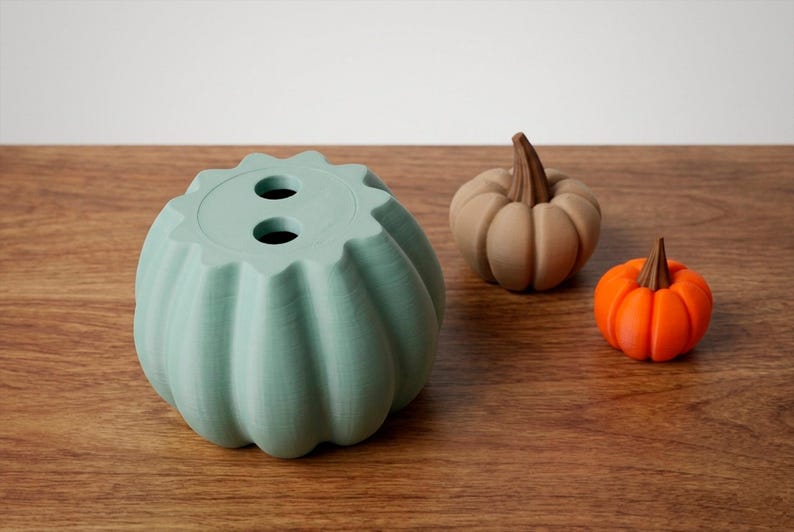 May include: A collection of decorative pumpkins in various sizes and colors. A large, light green pumpkin-shaped container with two holes sits next to a tan and an orange pumpkin. The pumpkins are on a wooden surface.