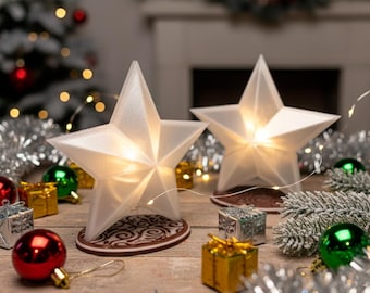 May include: Two illuminated white star-shaped Christmas decorations with a warm glow, set on decorative bases. The stars are surrounded by Christmas ornaments, including red and green baubles, silver tinsel, and small gift boxes, creating a festive holiday scene.