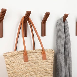 May include: Five brown wooden wall hooks with a ribbed design are mounted on a white wall. A woven straw tote bag with brown leather handles hangs from one hook, and a grey scarf hangs from another.