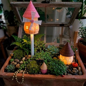 May include: A miniature fairy garden in a terracotta planter, featuring a windmill and a small house with glowing windows. The garden includes various plants, small stones, and a sign that reads "off-grid". A string of gold beads adds a decorative touch.