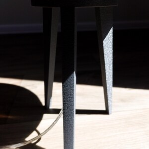 May include: Close-up of a dark gray metal table base with three legs. The legs have a textured surface and are angled slightly outward. The table's top is partially visible, with a light wood finish. A clear cord is visible.