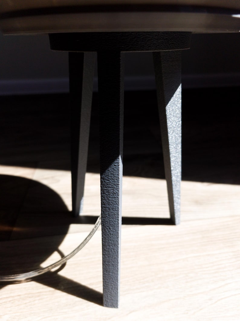 May include: Close-up of a dark gray metal table base with three legs. The legs have a textured surface and are angled slightly outward. The table's top is partially visible, with a light wood finish. A clear cord is visible.