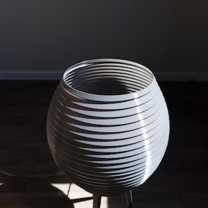 May include: A modern, gray, decorative lamp with a unique design. The lamp features a spherical shade constructed from layered, horizontal bands. It stands on a dark gray, three-legged base. The lamp is illuminated by natural light.