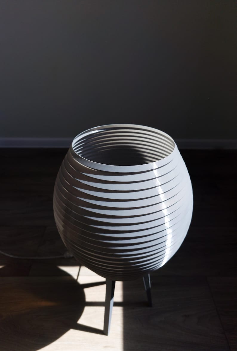 May include: A modern, gray, decorative lamp with a unique design. The lamp features a spherical shade constructed from layered, horizontal bands. It stands on a dark gray, three-legged base. The lamp is illuminated by natural light.