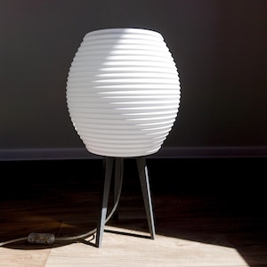 May include: White, ribbed, spherical lamp with a dark gray tripod base. The lamp has a textured surface and a cord with a clear plug. The lamp is on a wooden floor, with a dark background.