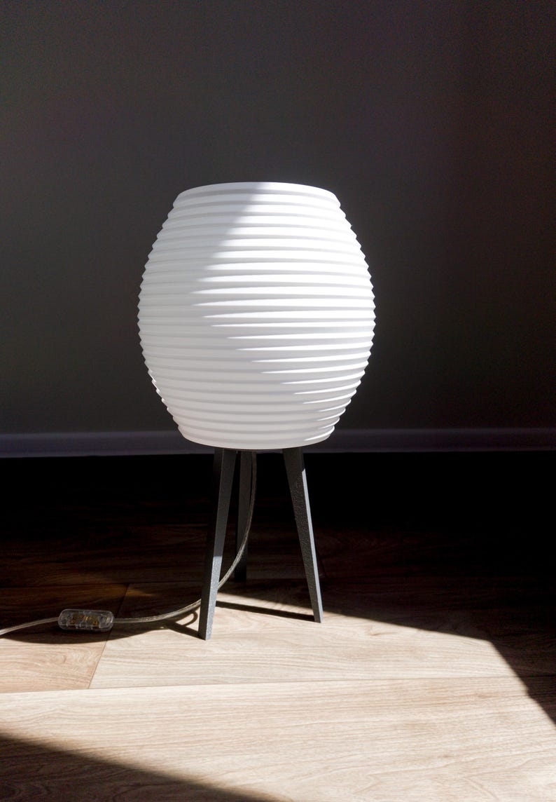 May include: White, ribbed, spherical lamp with a dark gray tripod base. The lamp has a textured surface and a cord with a clear plug. The lamp is on a wooden floor, with a dark background.