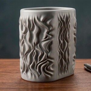 May include: A gray ceramic vase with a textured, flowing design. The rectangular vase sits on a wooden surface, with a silver pen resting nearby. The vase's surface has a rippled, organic pattern.