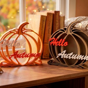 May include: Two decorative pumpkin-shaped ornaments made of layered material. The left pumpkin is orange with white and red text reading Hello Autumn. The right pumpkin is brown with red and white text reading Hello Autumn, displayed on a surface.