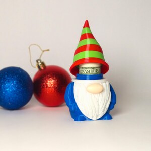 May include: A blue gnome figurine with a white beard and a red and green striped hat. The hat is lifted to reveal a rolled-up US dollar bill with the text OF AMERICA visible. Beside the gnome are one blue and one red glittery ornament.