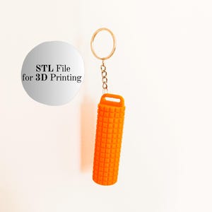 Puede incluir: Llavero cilíndrico naranja brillante y texturizado con un anillo y cadena dorados. El llavero tiene una abertura rectangular en la parte superior. Un círculo plateado con el texto "STL File for 3D Printing" está en el fondo.