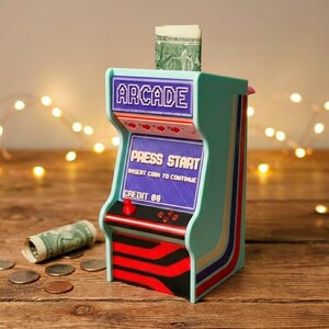 May include: A mint green arcade-style bank with red, black, and blue accents. The bank has a slot for inserting bills and a screen that reads "PRESS START". A rolled-up dollar bill and coins are on the wooden surface.