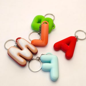May include: A collection of five letter-shaped keychains featuring the letters M, L, A, F, and B. The keychains are made of smooth, rounded plastic in various colours including red, orange, light blue, lime green, and a tan letter M with white.