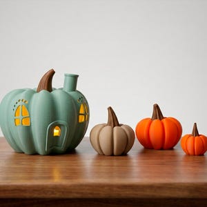 May include: A collection of decorative pumpkin figurines in various sizes and colors. The largest is a teal pumpkin-shaped house with windows and a chimney. Other pumpkins are tan and orange, with brown stems. The pumpkins are displayed on a wooden surface.