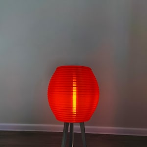 May include: A vibrant red, ribbed lamp with a cylindrical shape, illuminated from within. The lamp sits on a gray, three-legged stand. The light glows brightly, casting a warm ambiance. The lamp's design is modern and eye-catching.