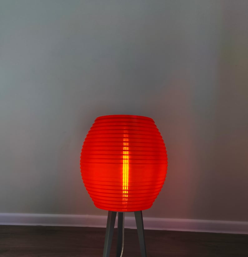 May include: A vibrant red, ribbed lamp with a cylindrical shape, illuminated from within. The lamp sits on a gray, three-legged stand. The light glows brightly, casting a warm ambiance. The lamp's design is modern and eye-catching.