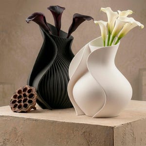 Op de afbeelding: Twee decoratieve vazen, een zwarte en een witte, elk met bloemen. De zwarte vaas heeft een gestructureerd, geribbeld ontwerp en bevat donkerpaarse calla lelies. De witte vaas bevat witte calla lelies. Een gedroogde zaaddoos ligt ervoor.