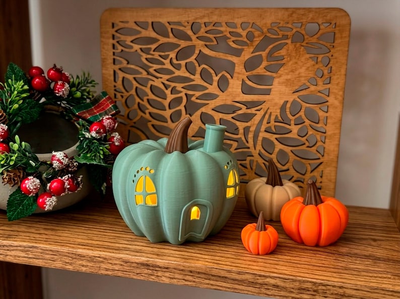 May include: A teal pumpkin-shaped house with illuminated windows and a brown stem sits on a wooden shelf. Beside it are three pumpkins in orange and cream colors. A decorative wreath with red berries and a wooden tree cutout complete the autumn-themed display.