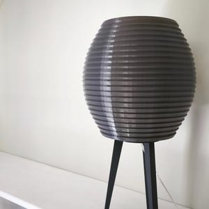 May include: A gray, ribbed, spherical lamp shade sits atop a black tripod base. The lamp is on a white shelf against a white wall. The lamp's shade has a series of horizontal lines, creating a textured appearance.