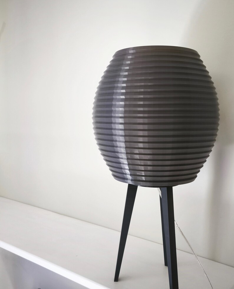 May include: A gray, ribbed, spherical lamp shade sits atop a black tripod base. The lamp is on a white shelf against a white wall. The lamp's shade has a series of horizontal lines, creating a textured appearance.