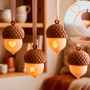 May include: Four acorn-shaped hanging ornaments with brown textured caps and glowing light-colored bodies. Three ornaments feature cutouts of a heart, a star, and a leaf. An inset shows one acorn hanging from a curved stand on a flat base.