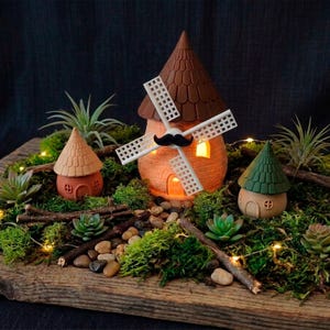 May include: A miniature fairy garden scene featuring a windmill with a mustache, two small houses, succulents, moss, and decorative lights. The windmill and houses are made of terracotta-colored material with brown and green accents, set on a wooden base.