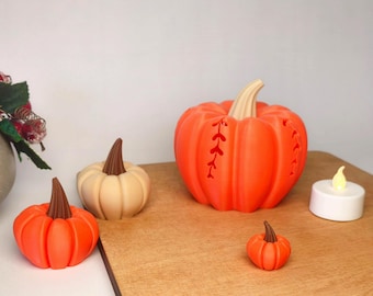 Puede incluir: Un conjunto de cuatro calabazas decorativas de varios tamaños y colores, incluyendo dos calabazas de color naranja brillante, una calabaza beige y una pequeña calabaza naranja, todas con tallos marrones, junto a una pequeña vela blanca de té.